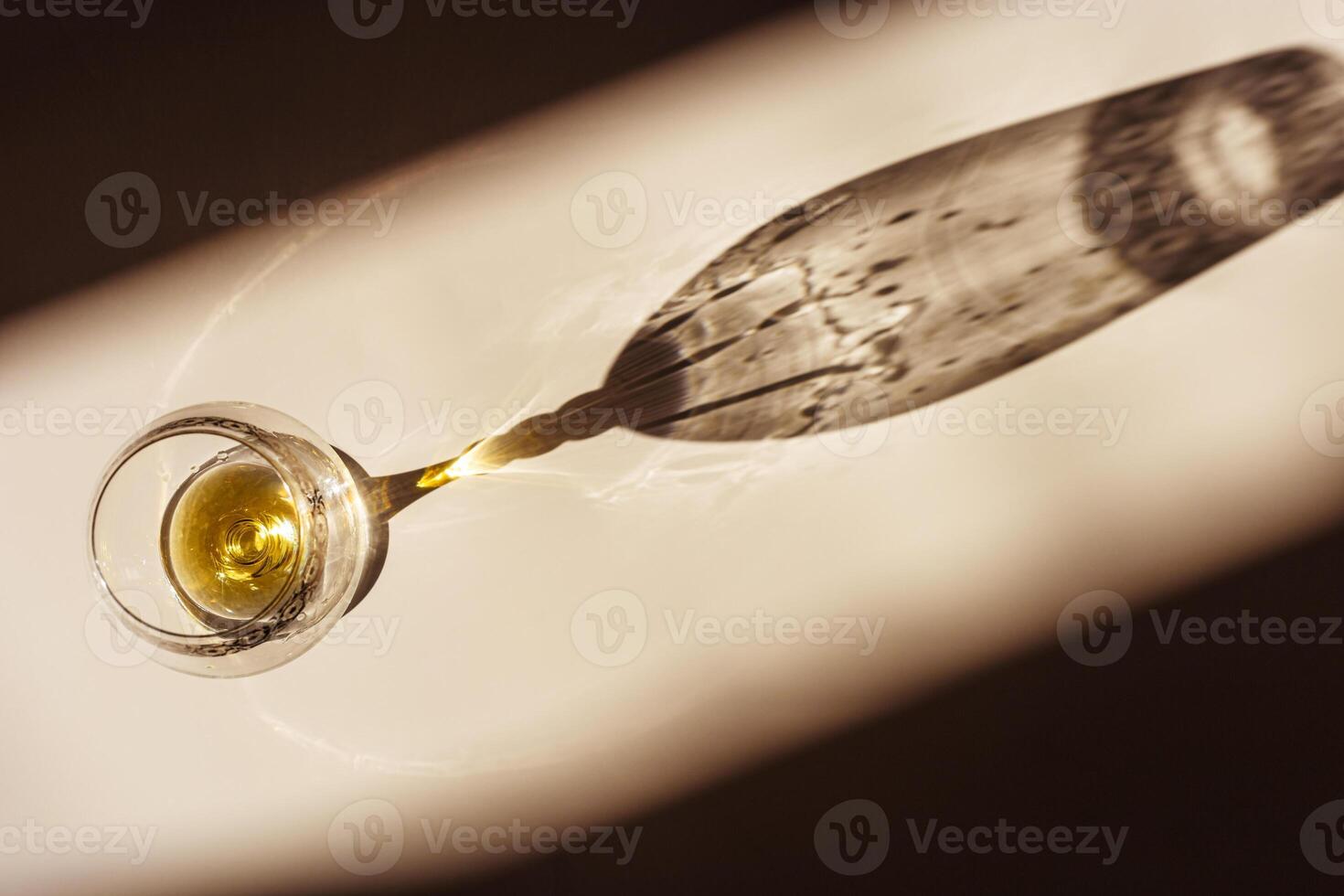 One glass of cognac sparkling at sunlight, beige background with dark shadow and reflection, sun flare light effect on table. Strong alcohol drink with star filter, top view, desaturated colors photo