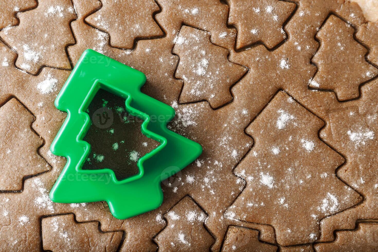 Cooking ginger cookies, Christmas New Year traditional cookies in shape of Christmas fir tree, raw rolled out dough with cut out dough figures, closeup, top view, green plastic pastry cutter on dough photo