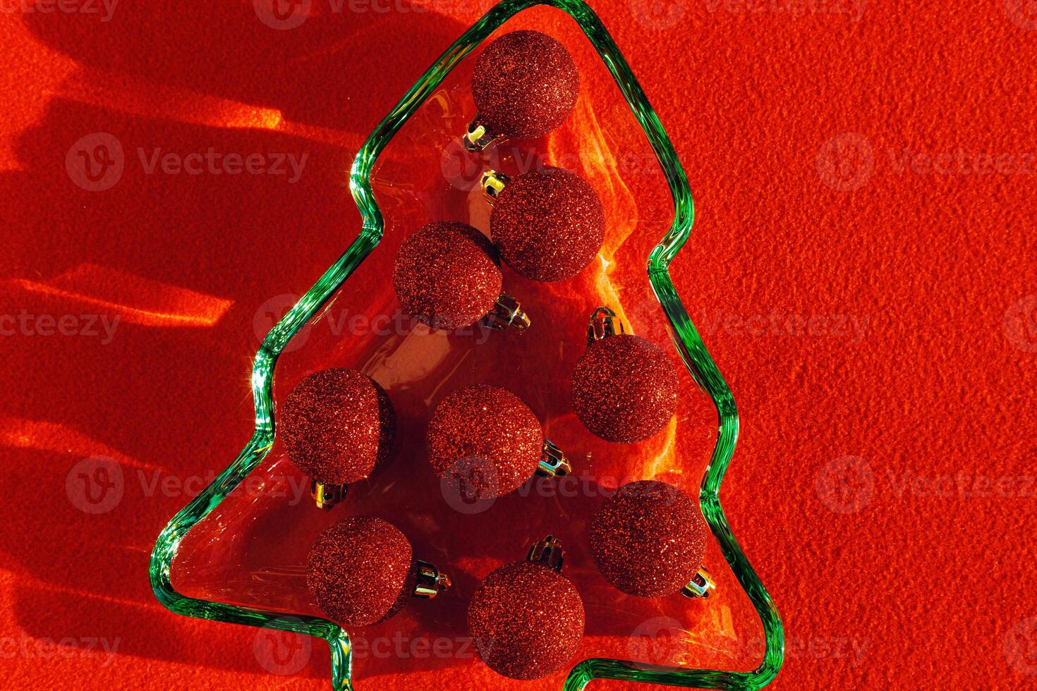 Green colored glass plate form as New Year tree with sparkling red Christmas balls instead of food, sunlight, shadow, star glare on red cloth background. Winter holiday dinner concept, minimal photo