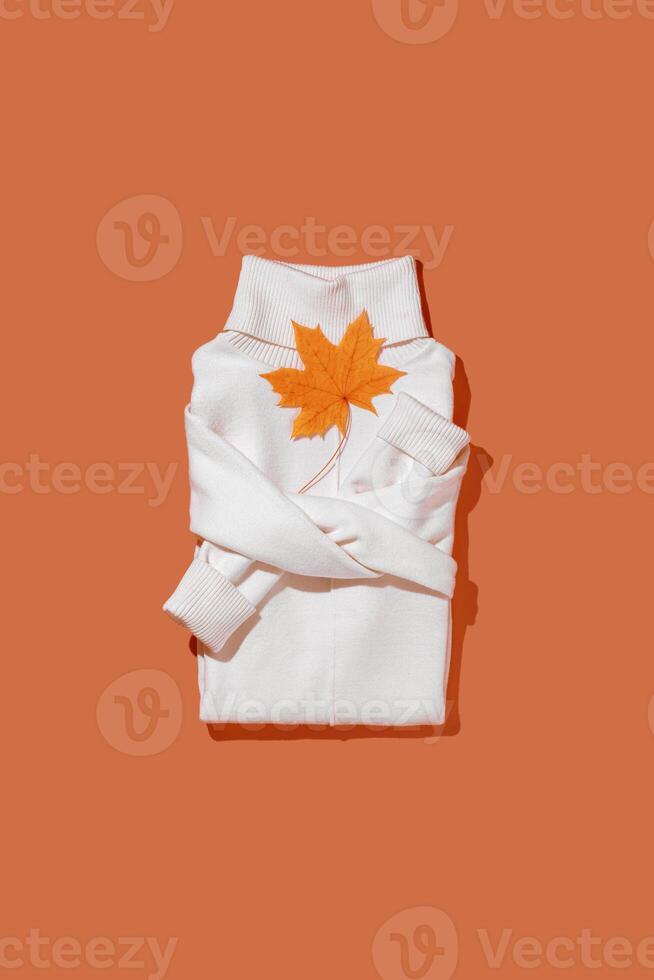 White warm folded sweater with autumn maple leaves on vivid orange color background, folded women jumper top view, minimal style trend autumn clothing concept, cozy clothes Aesthetic vertical Flatly photo