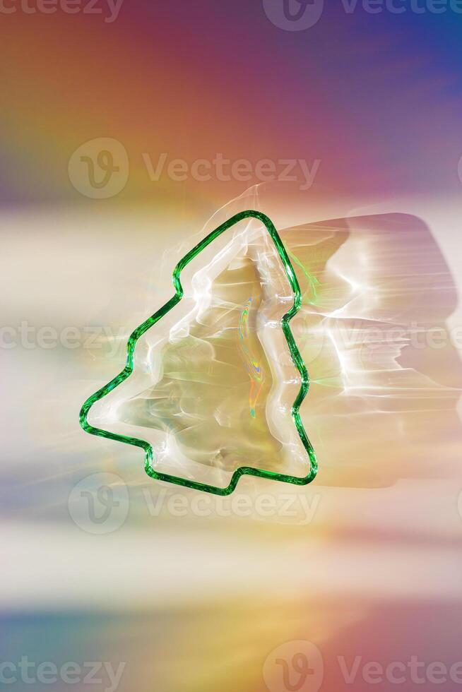 Green colored glass form as New Year tree, Christmas fir at sunlight with beautiful shadows and sparkling sun glare on rainbow gradient background. Winter holiday concept, minimal aesthetic photo