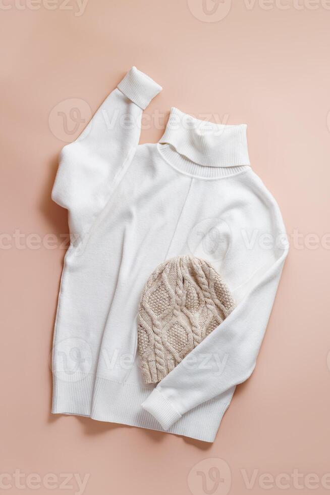 White turtleneck or sweater and knitted hat, casual garment, comfort and style warm clothes. Soft pullover and wool hat on beige pink color background, minimal fashion clothing, top view photo