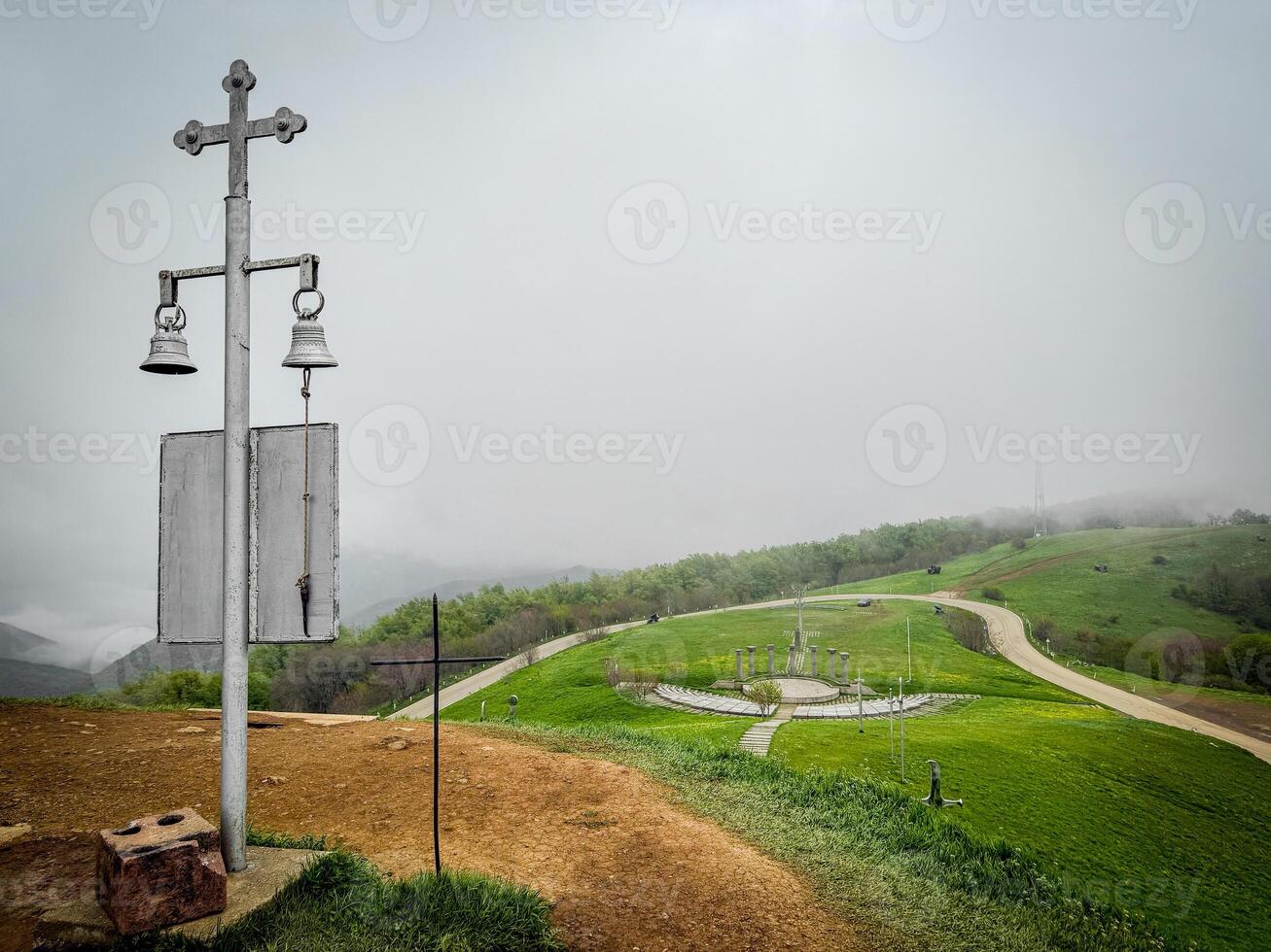 Georgia, Didgori, Sakartvelo. Metal bells and a cross rise from a misty hilltop, Metal bells and a cross rise from a misty hilltop, marking a tranquil spiritual monument along a winding rural pathway photo
