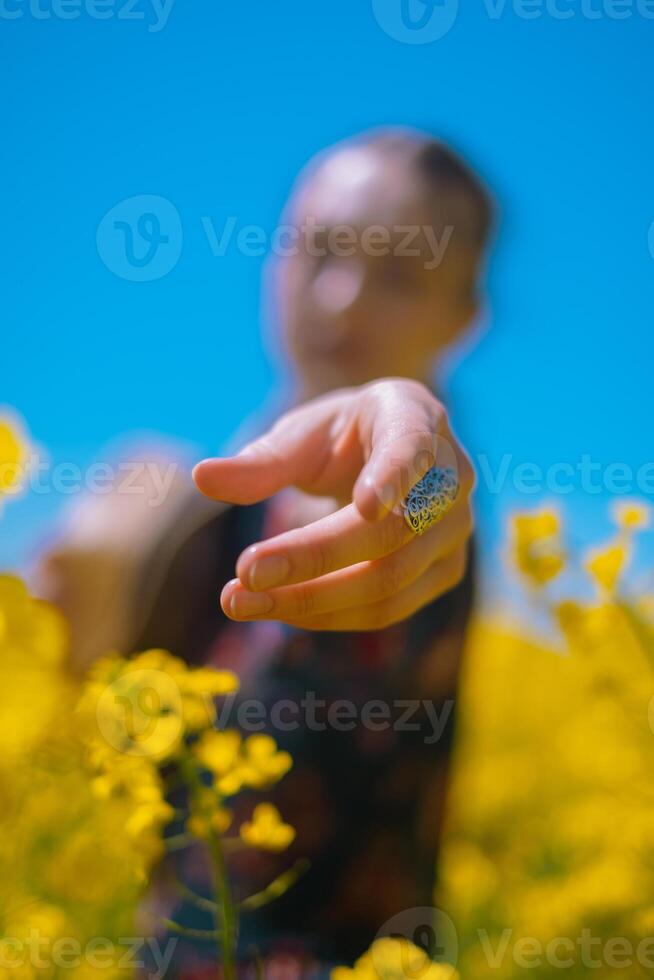 Close-up of girl reaching towards camera with silver jewelry on her arm, holding handcrafted round straw bag in blooming rapeseed field. photo
