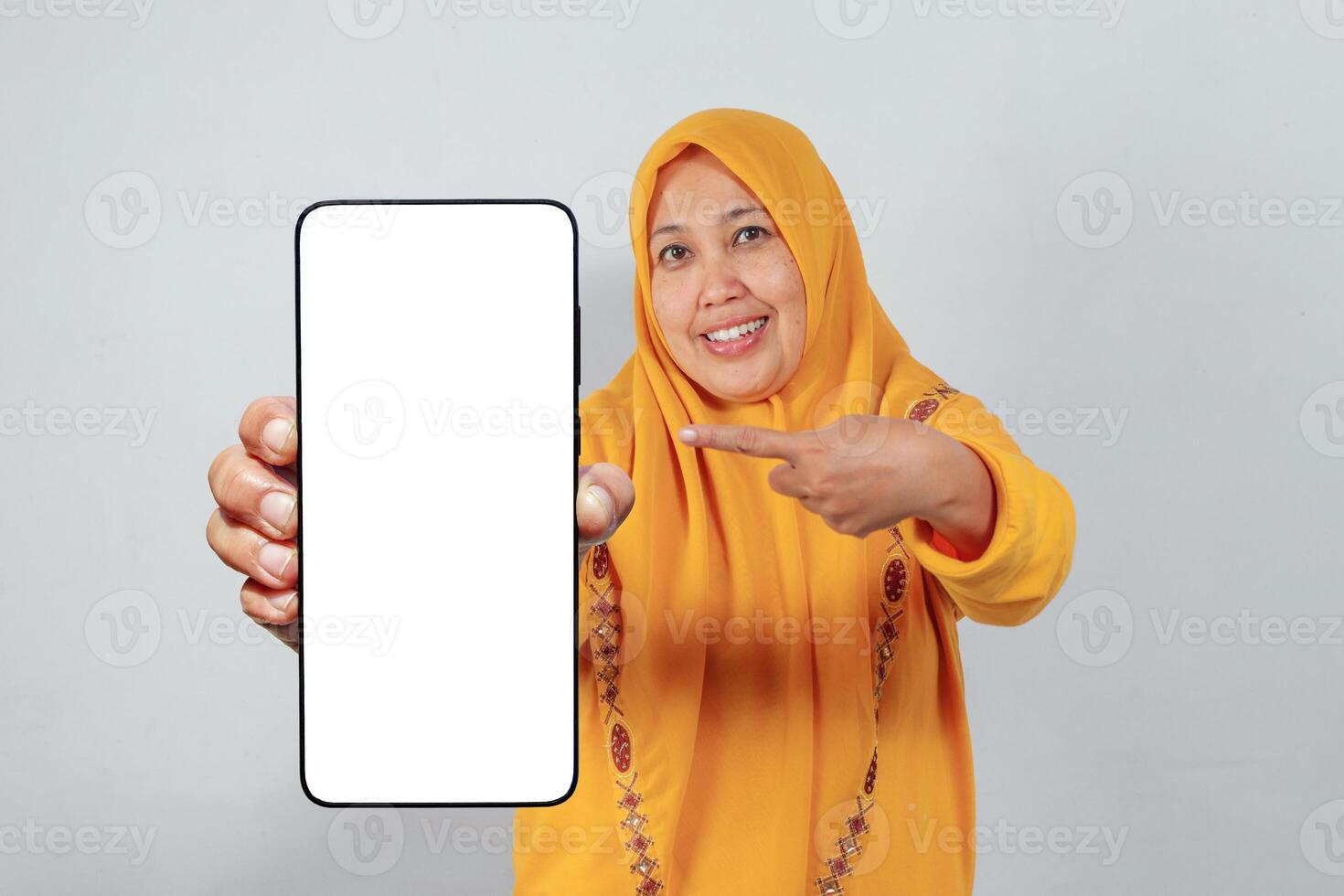 Muslim woman in yellow hijab smiling and pointing at smartphone with blank screen photo