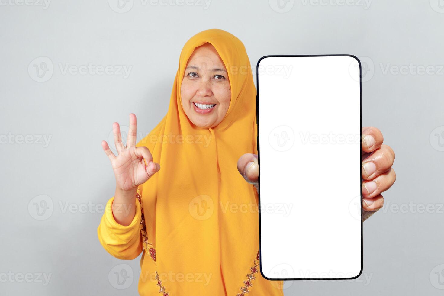 Woman in yellow hijab smiling while showing phone screen and making OK gesture photo