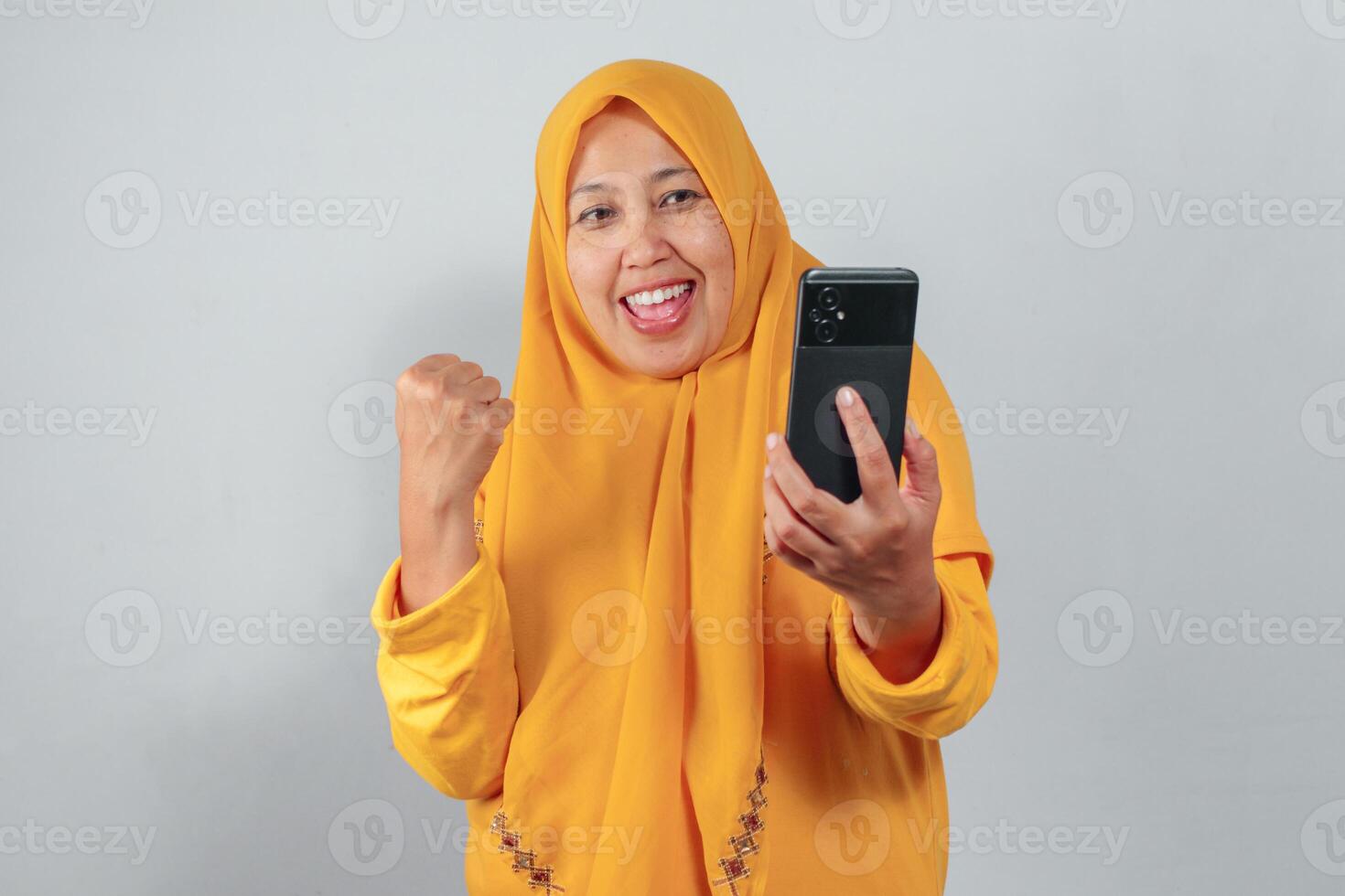 Muslim woman in yellow hijab cheering with fist up while looking at smartphone happily photo