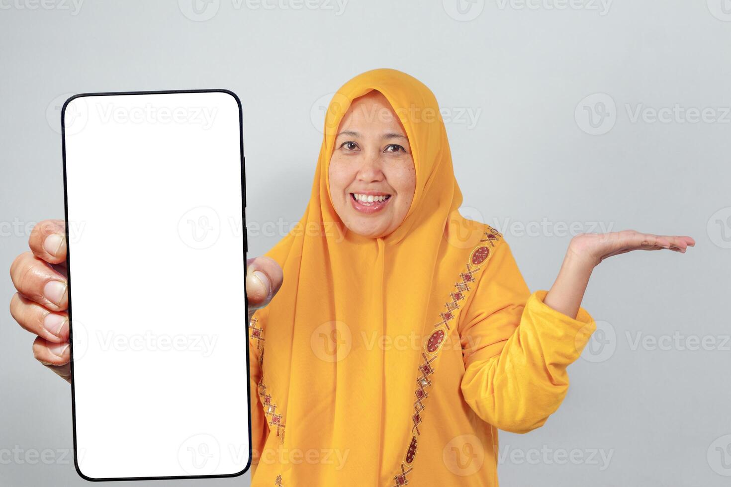 Smiling woman in yellow hijab showing phone screen and presenting with open hand photo