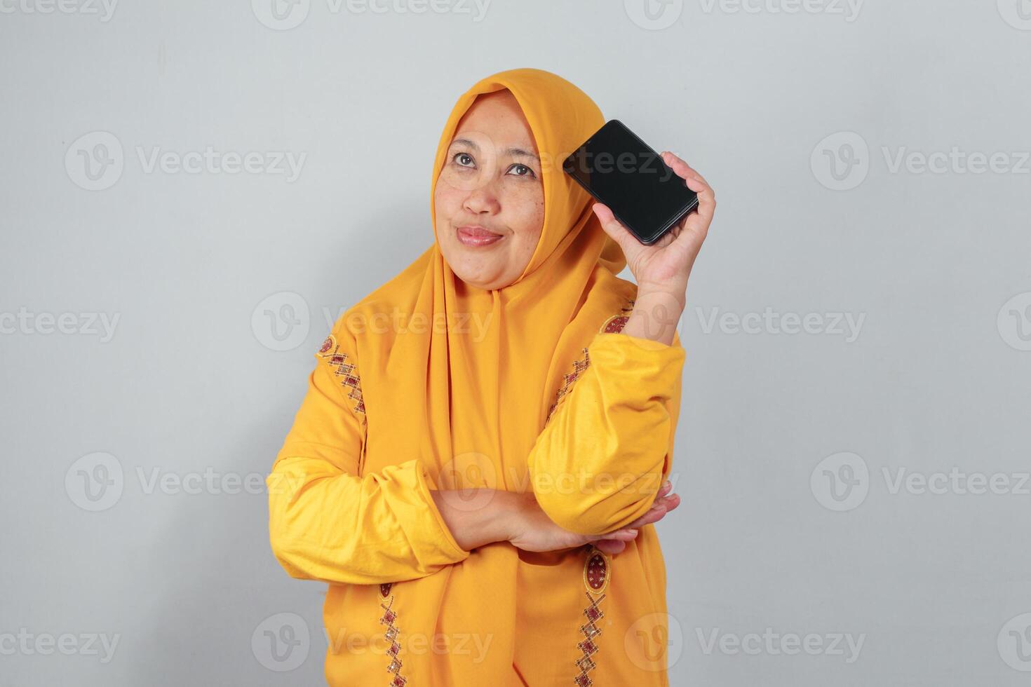 Middle aged woman in yellow hijab holding phone near head and looking up thoughtfully photo