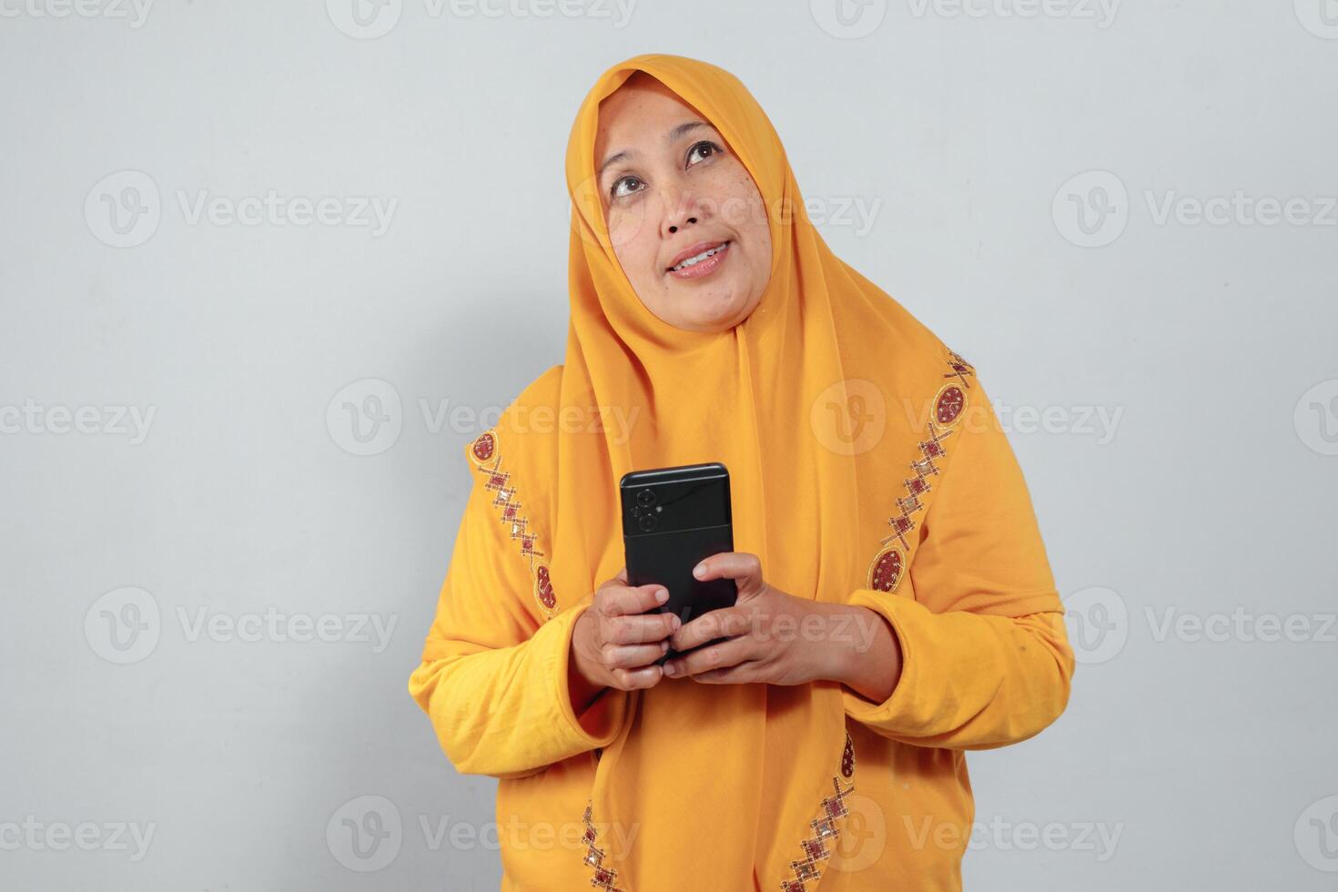 Muslim woman in yellow hijab holding phone with both hands and looking up thoughtfully photo