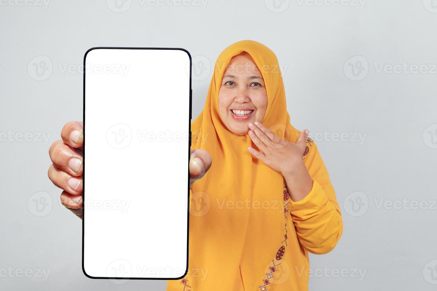 Smiling woman in yellow hijab holding blank phone and reacting with surprise gesture photo