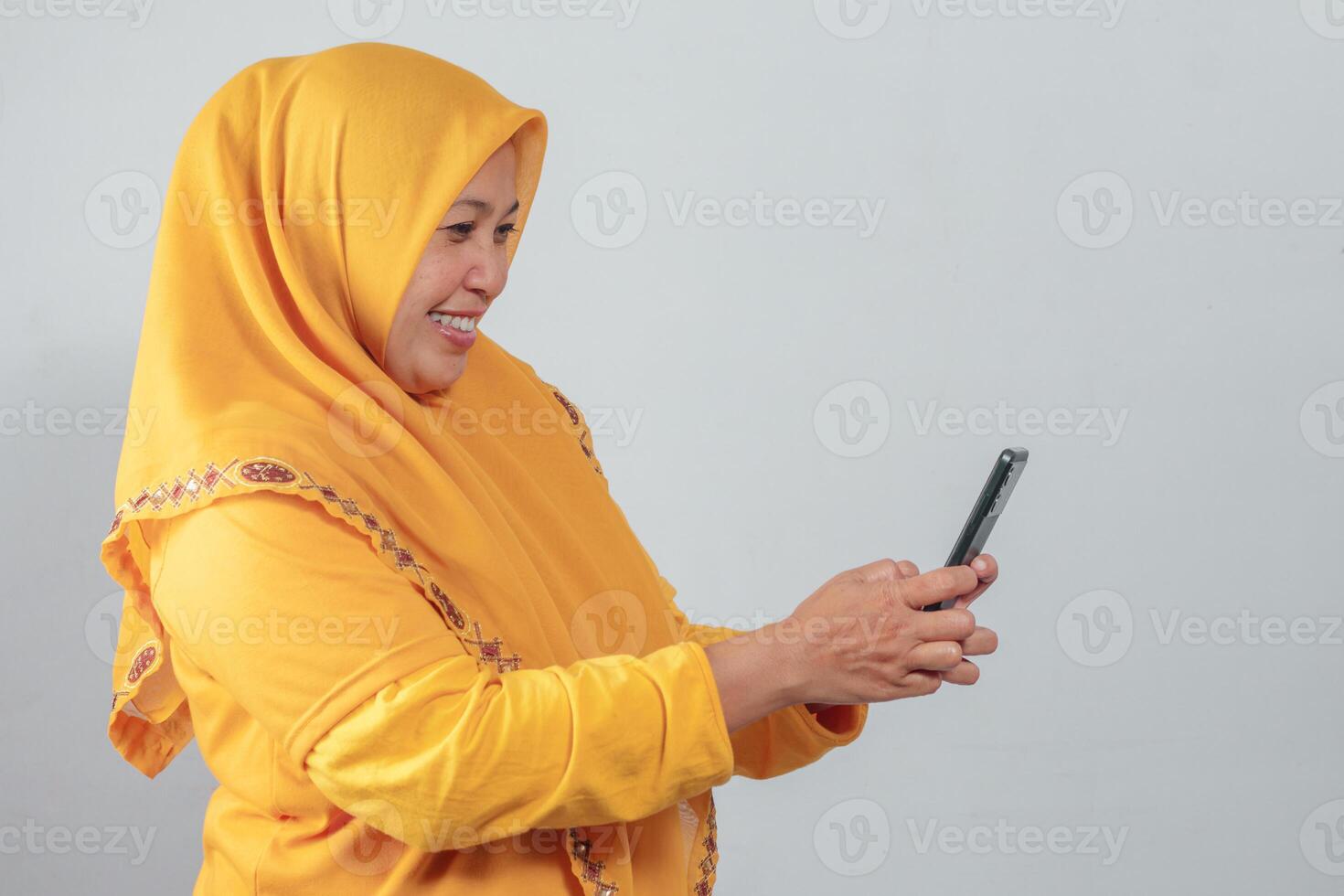 Muslim woman in yellow hijab smiling and using smartphone with both hands in side view photo