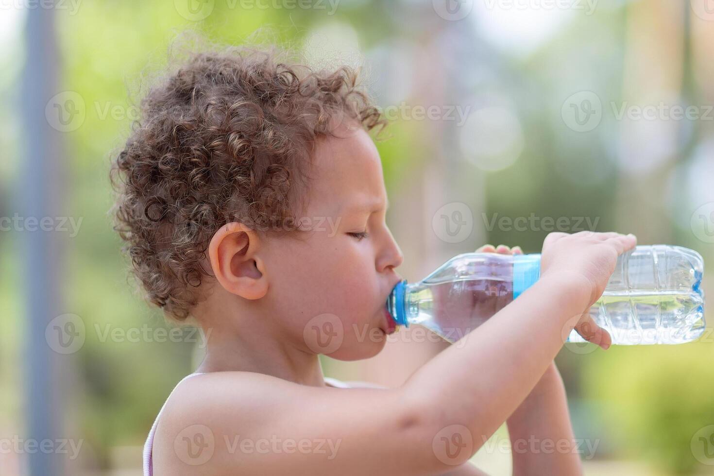 A curly child drinks water from a plastic bottle on a hot day photo