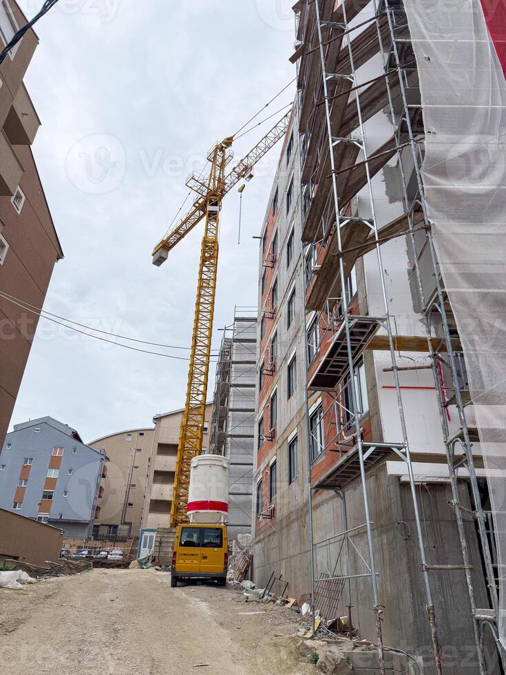 Tall construction crane beside unfinished apartment block. The building is surrounded by scaffolding, with construction materials and a dirt road below. photo