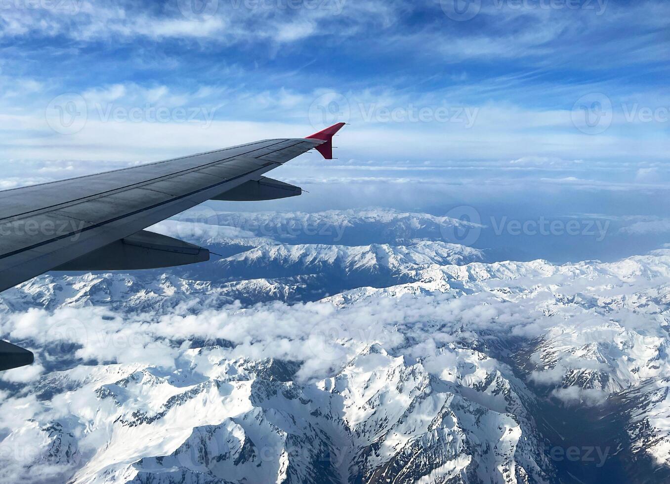 stunning aerial view of rugged snow-covered mountain peaks stretching into the horizon, viewed from an airplane window with the wing partially visible, under vivid blue sky with high-altitude clouds photo