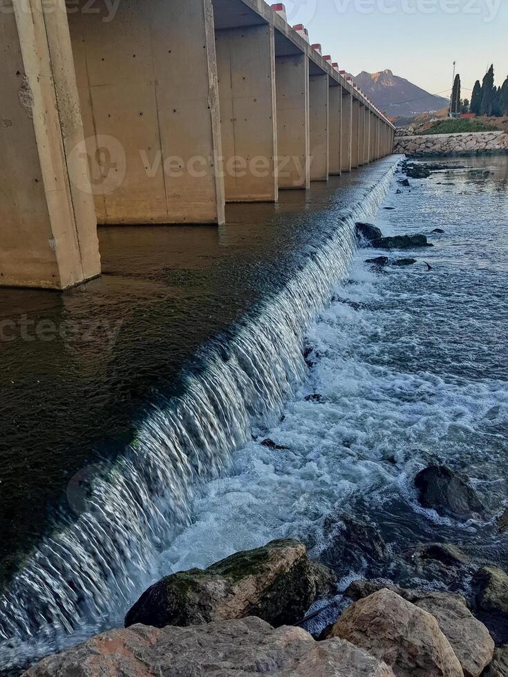 A wide waterfall cascades beneath a bridge in the heart of a Wadi. As you approach the flowing water, intricate details emerge, unveiling dynamic patterns and textures. photo