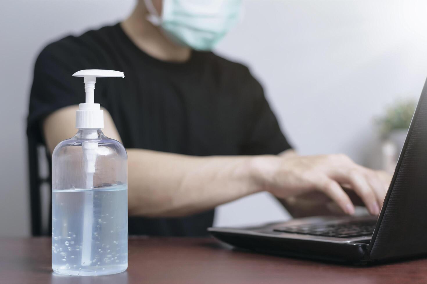 Adult man working remotely at home with laptop, wearing a face mask and using hand sanitizer for hygiene and virus prevention. Concept of online job, healthcare, and safe lifestyle during outbreak photo