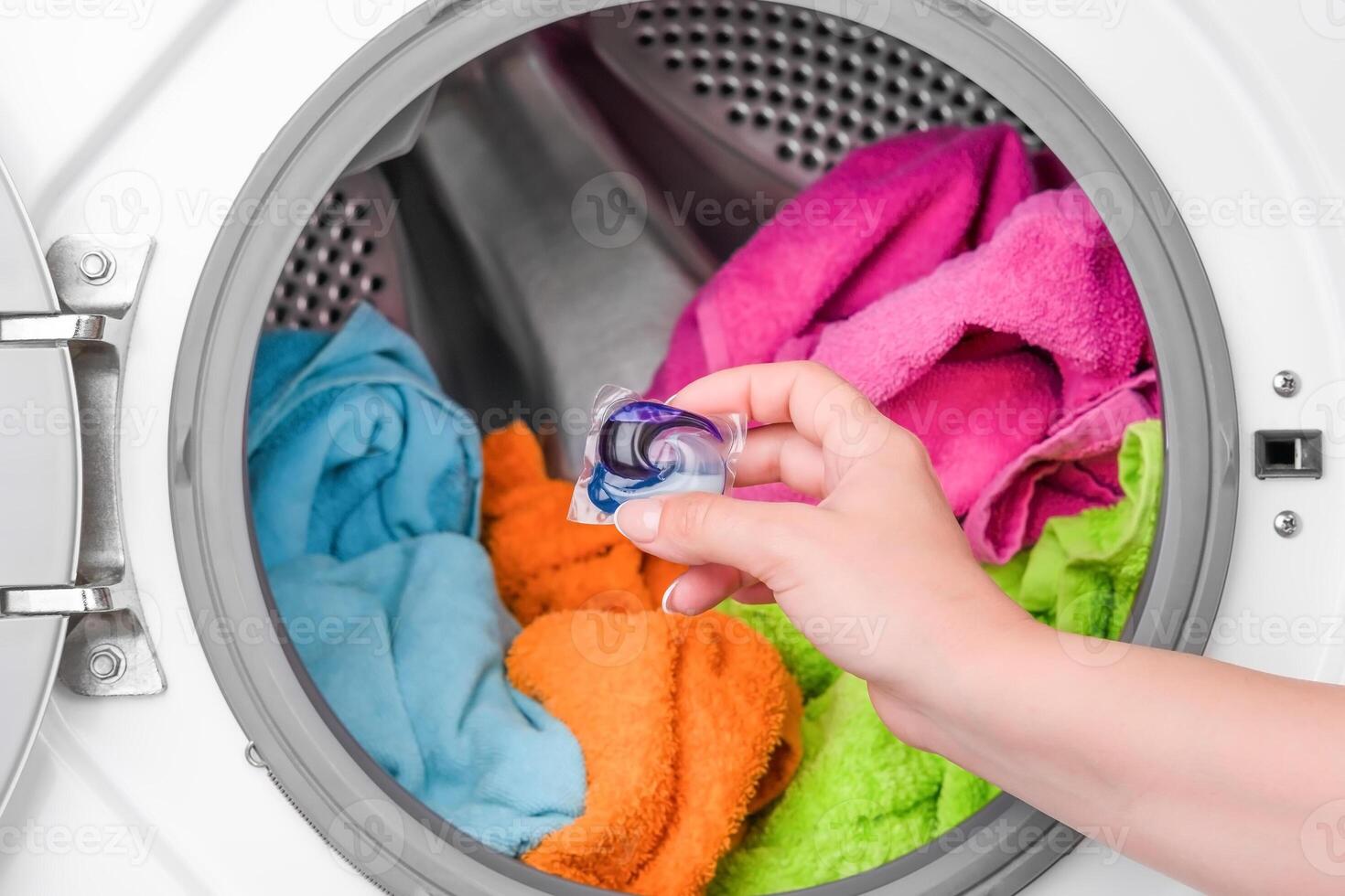 female hand holding washing capsule against background of washing machine with colored laundry photo