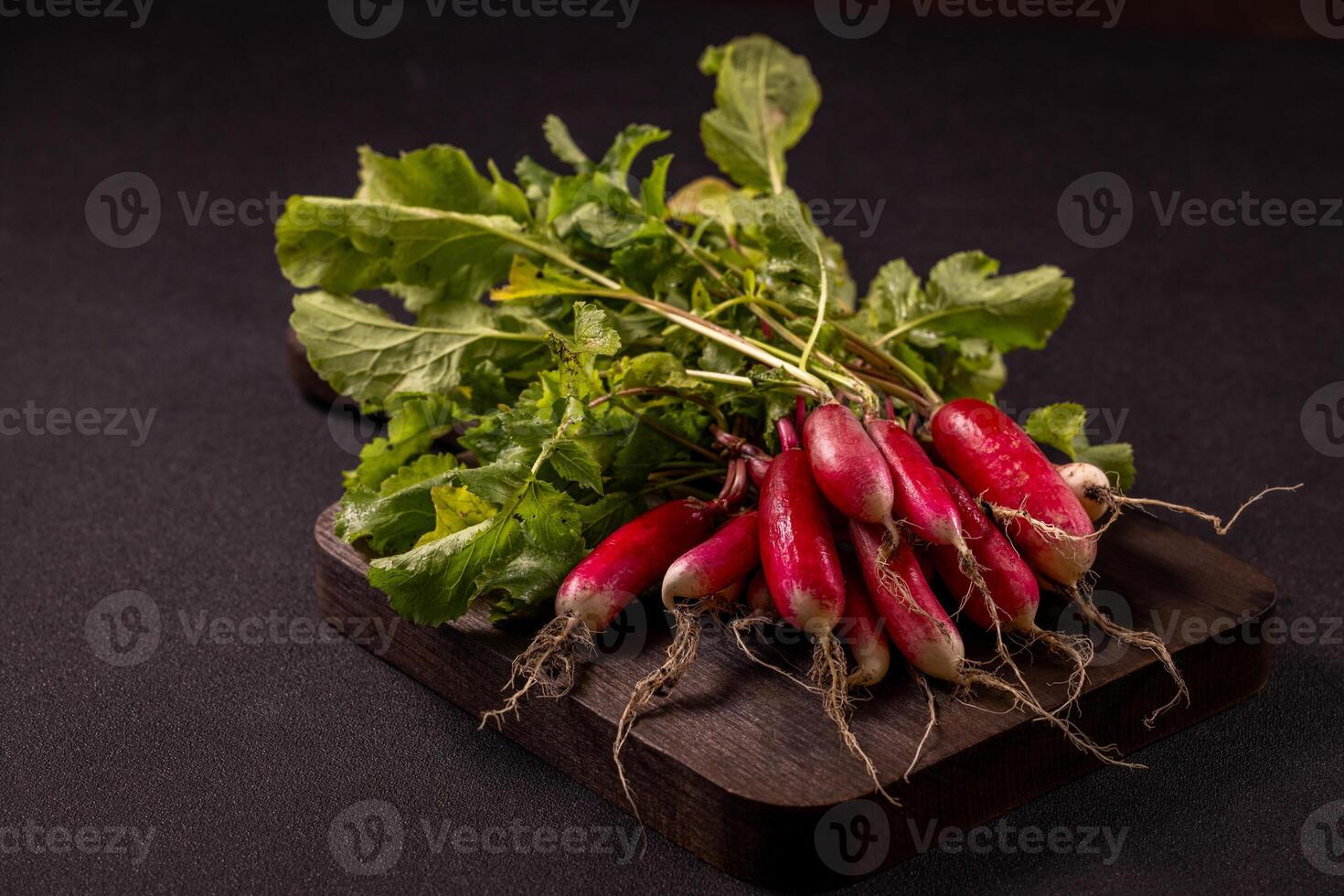 Vibrant red radishes with lush green leaves sit on a dark wooden cutting board, creating a simple yet appealing display of fresh produce photo