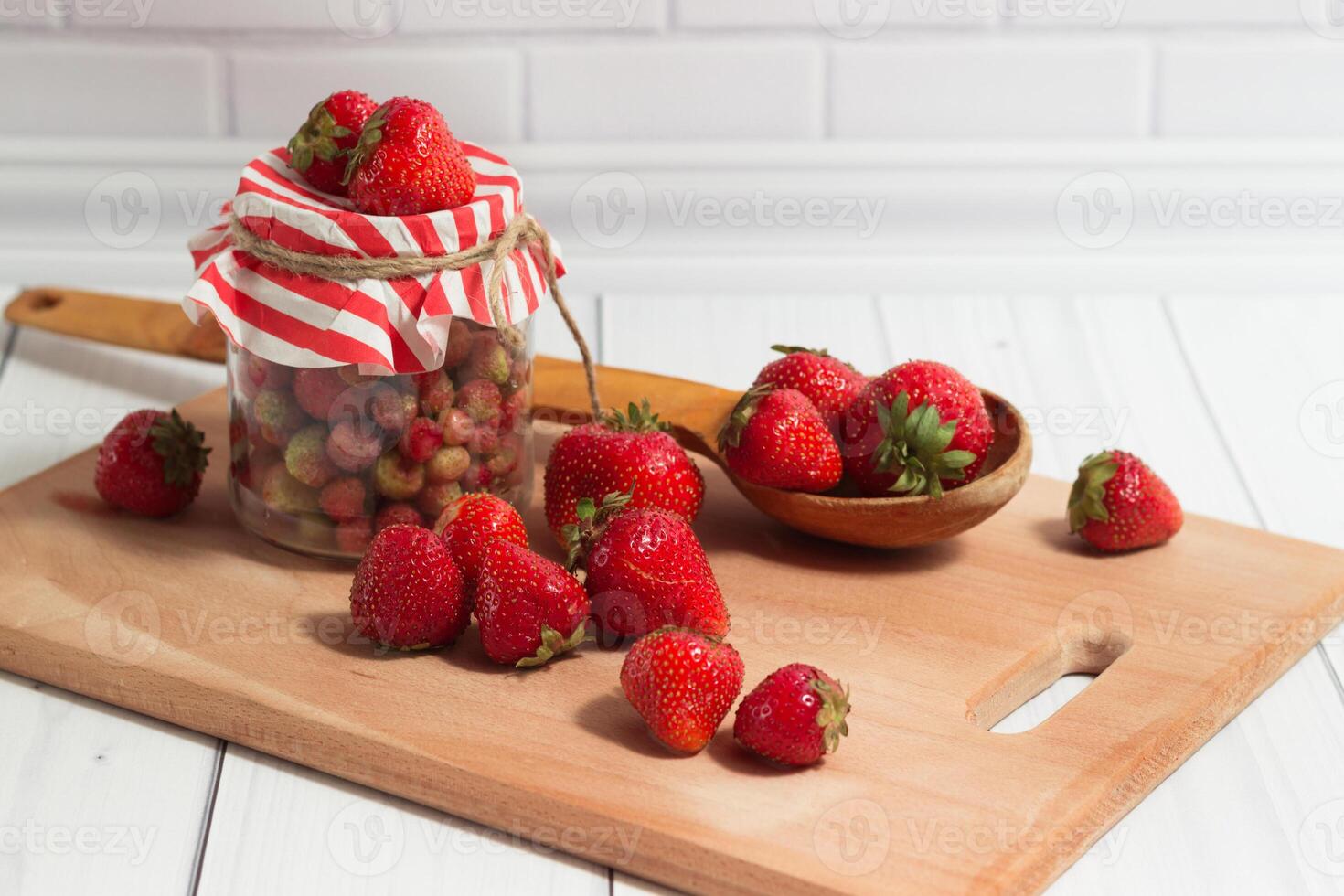 Ripe red strawberry in the glass jar with striped top and wooden spoon on the board on the light gray background photo