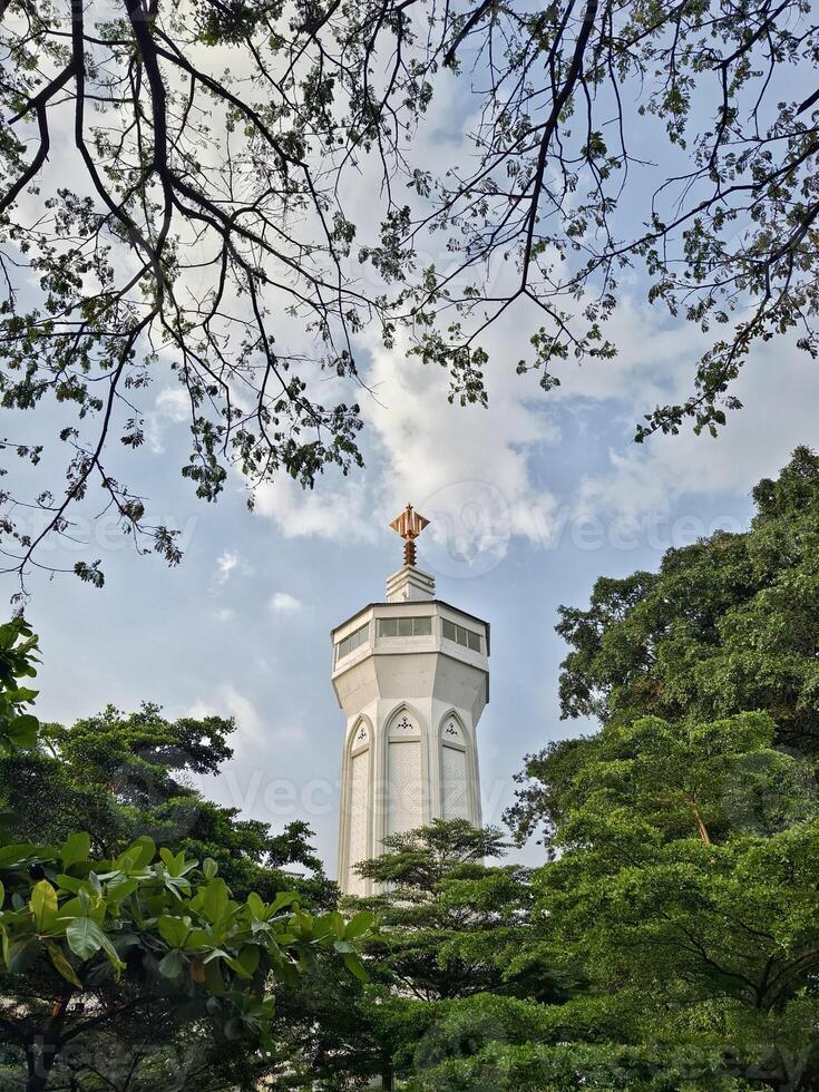 A white mosque minaret with traditional Islamic architectural details and a golden top, framed by green tree branches and blue sky photo