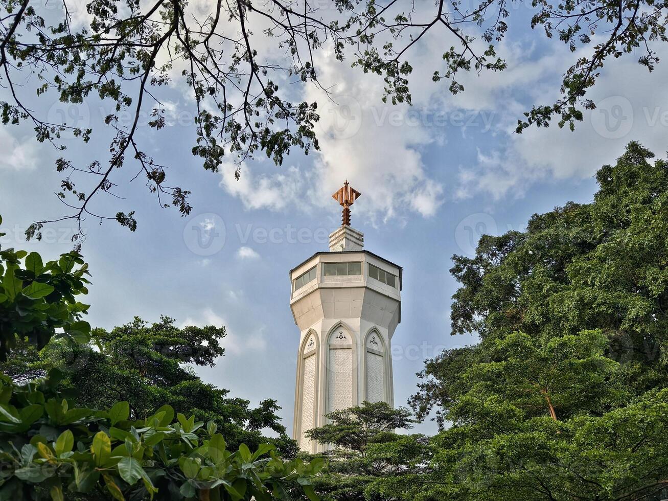 A white mosque minaret with traditional Islamic architectural details and a golden top, framed by green tree branches and blue sky. photo