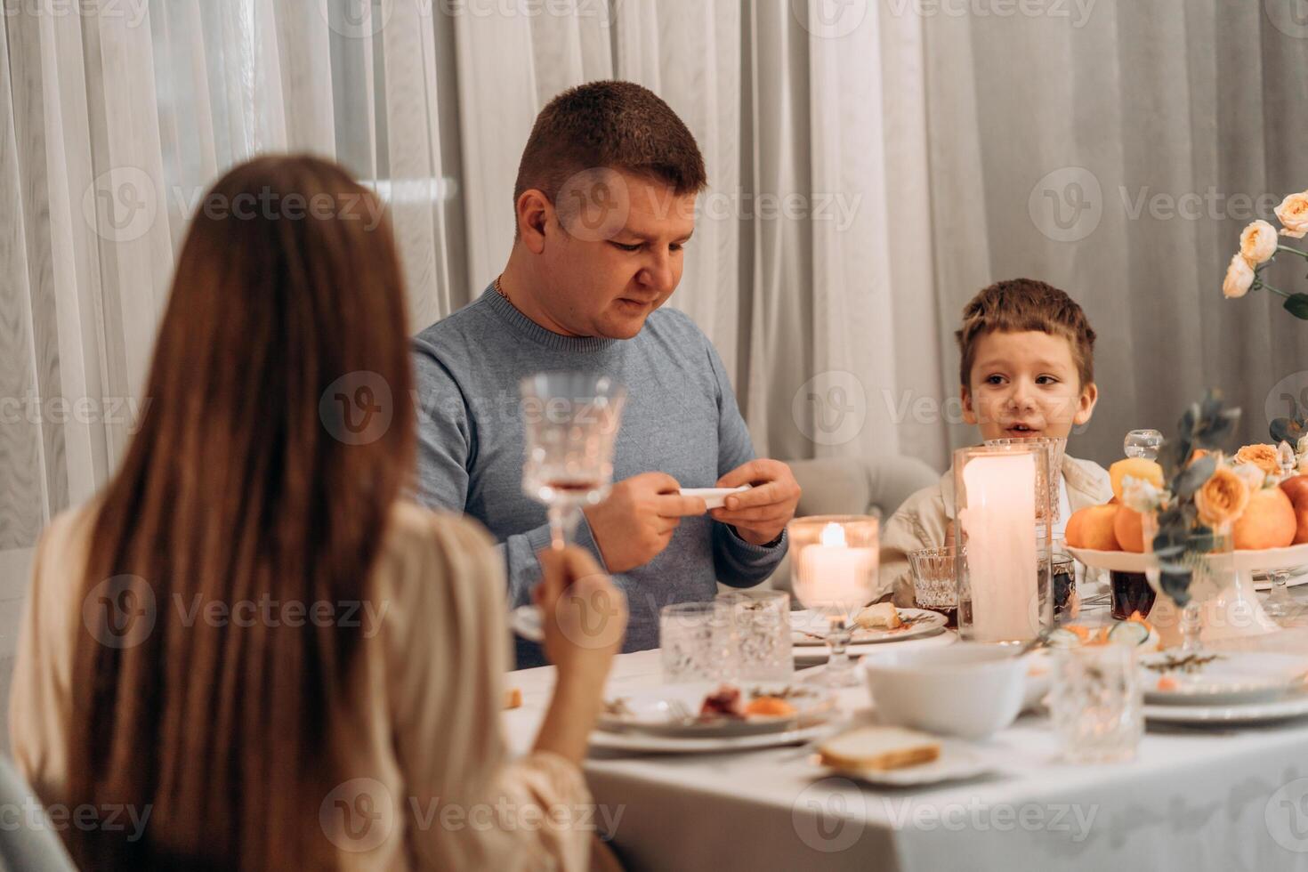 Father and son share thoughtful dinner moment at cozy table with candles fruit and flowers while mother listens from foreground capturing authentic family interaction warmth connection and mindful photo