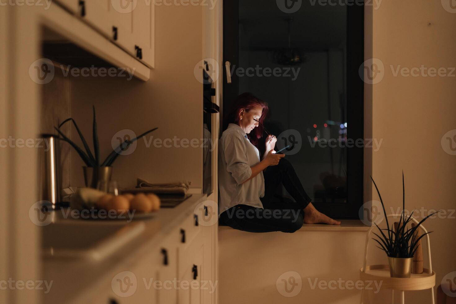 Woman sits in windowlight alone at night looking at glowing smartphone representing blue light at night digital intimacy and emotional solitude during evening scrolling routine in modern lifestyle photo
