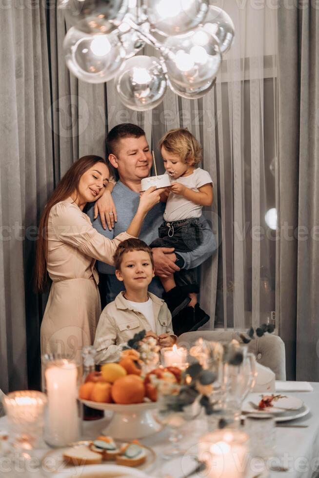 Smiling family gathered festive dinner table father holds toddler eating white cake mother lovingly supports child and older sibling stands nearby capturing heartwarming moment connection celebration photo