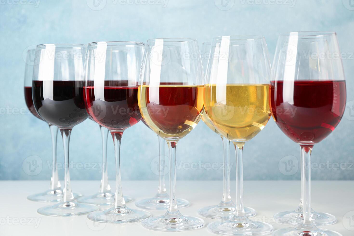 Glasses with different wine on white table against light blue background photo