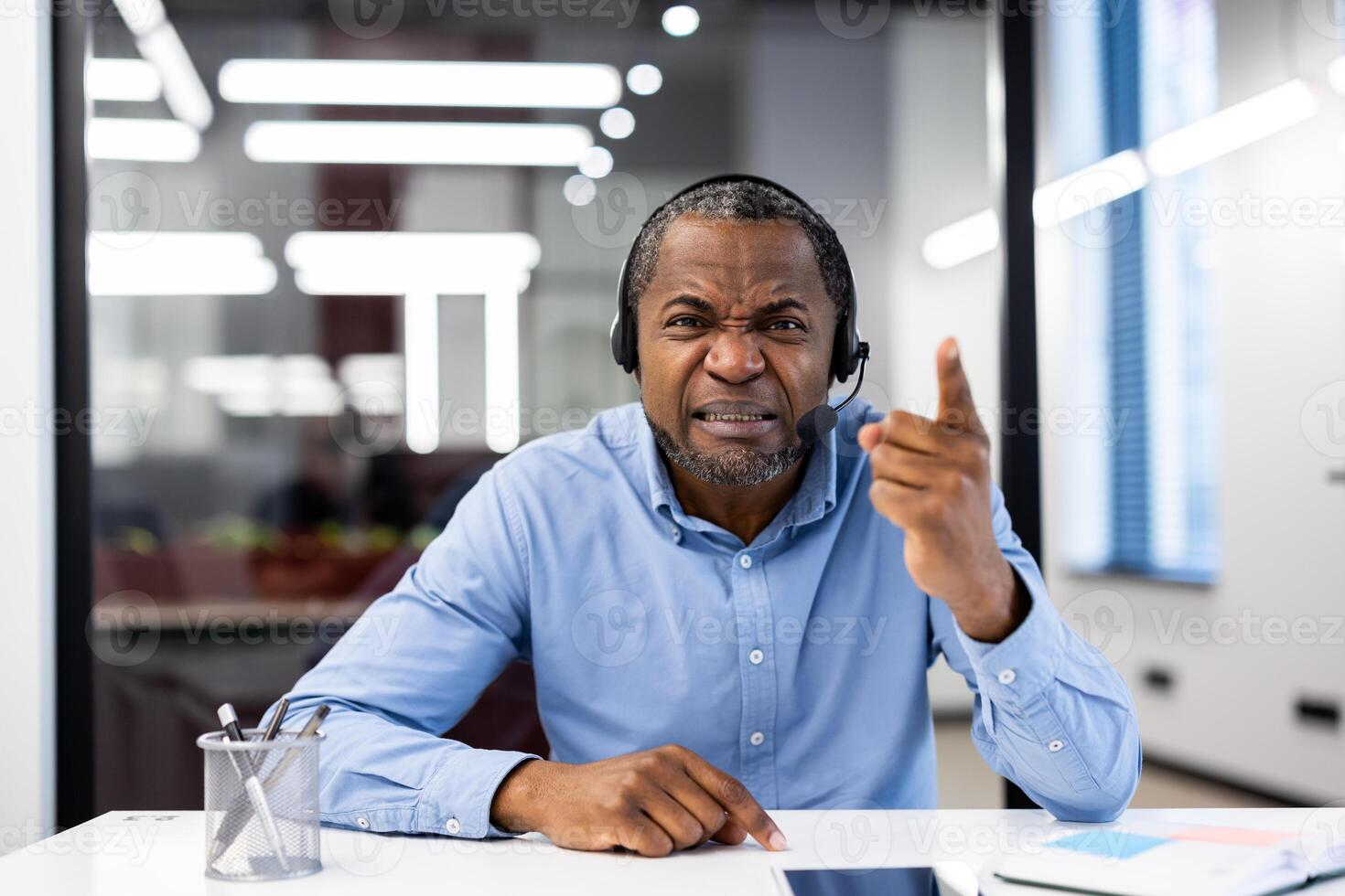 Mature african american businessman wearing headset expresses frustration in office. Engaged in intense phone call, pointing finger during a virtual meeting. Concept of leadership, communication. photo