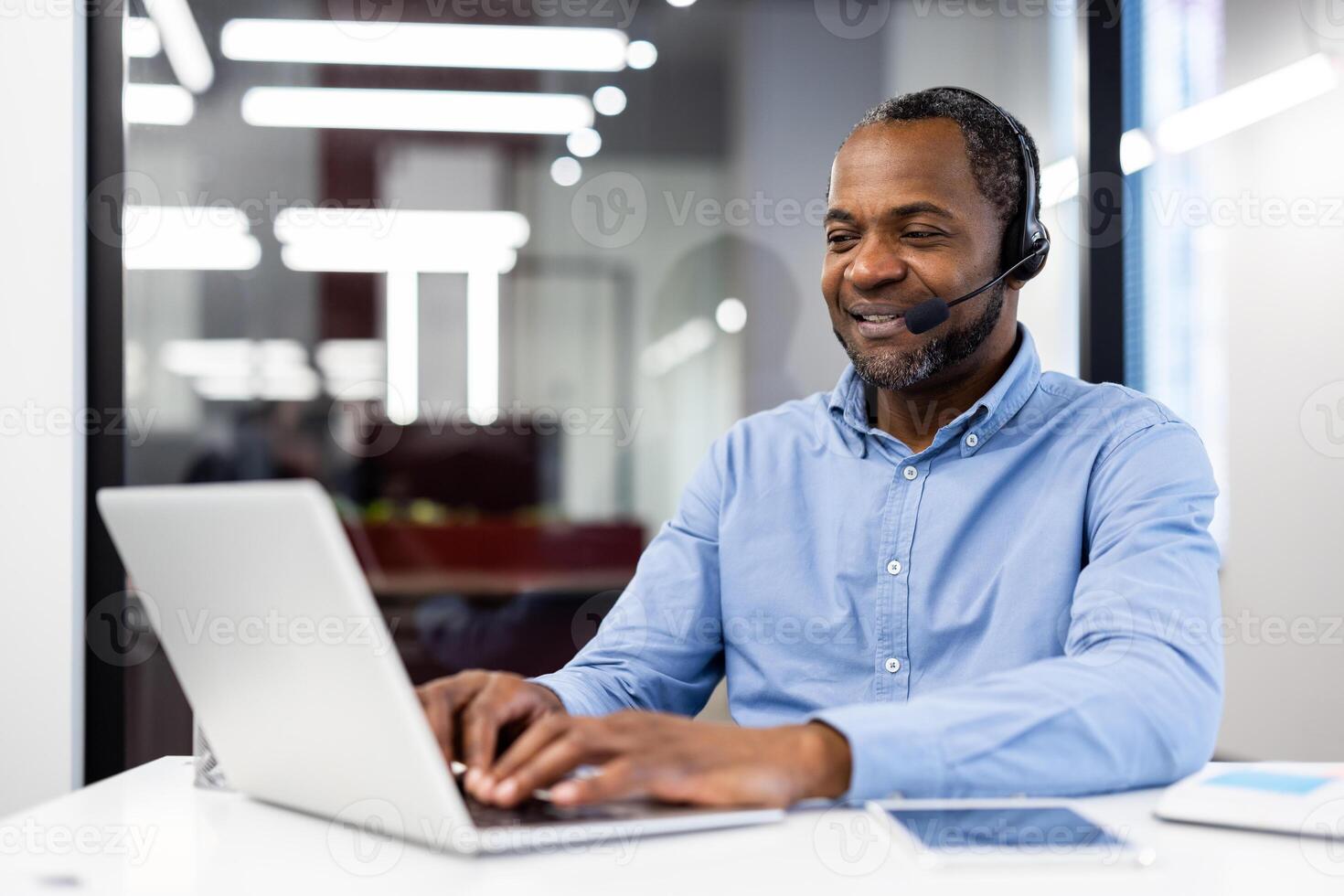 Mature african american businessman in modern office setting using laptop and headset. Displaying professionalism, communication skills, and confidence while typing and engaging with clients. photo
