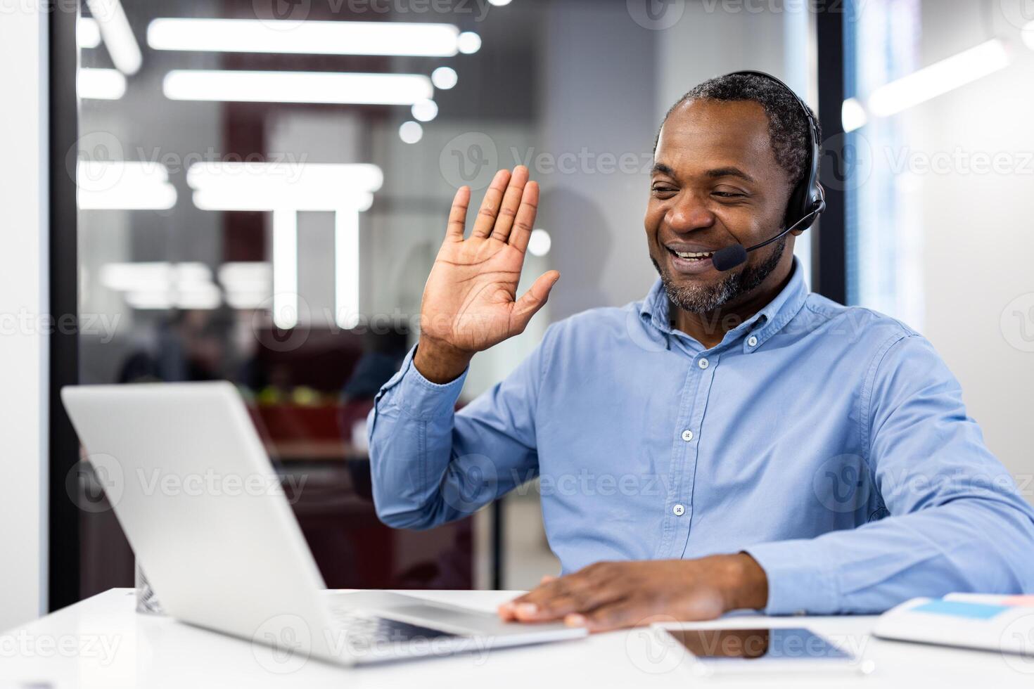 Mature african american male businessman wearing headset smiling while waving at laptop during call. Engaged in online meeting in modern office setting. Concept of communication, technology. photo