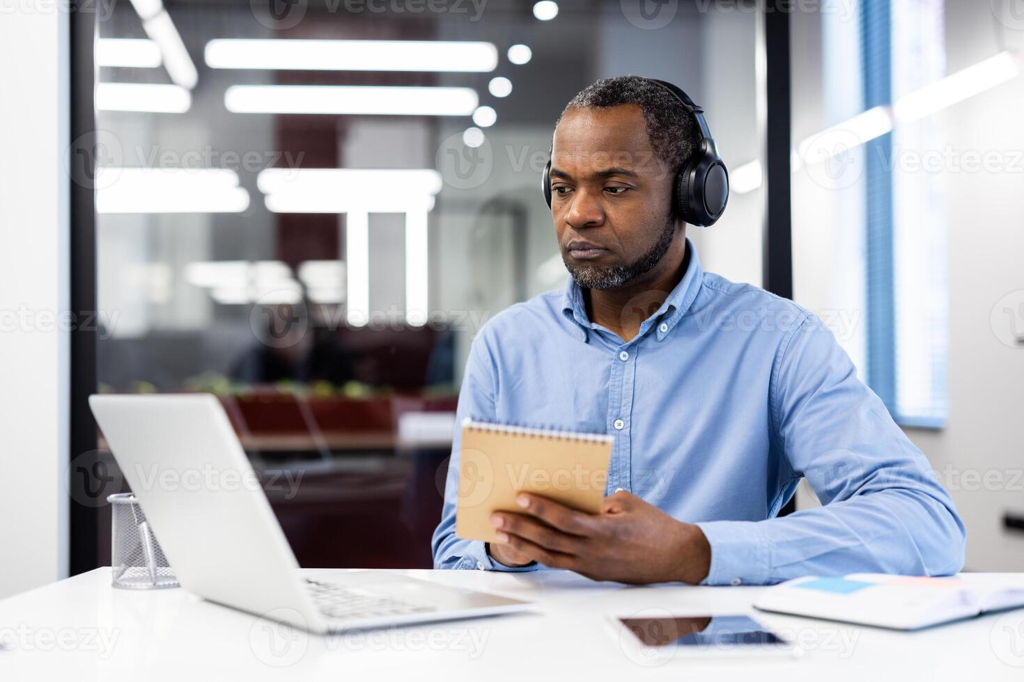 Mature African American businessman wearing headphones, working intently at desk in modern office setting. Holding notebook, engaging in online meeting via laptop, illustrating business focus photo