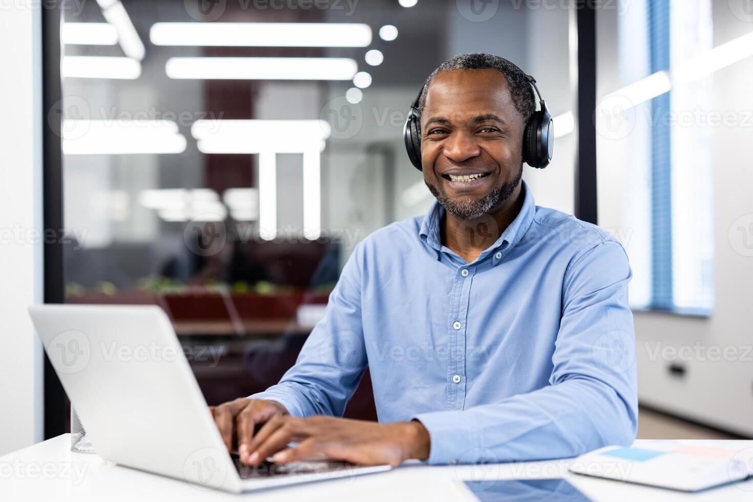 Mature African American businessman smiling while using laptop in office. Wearing headphones suggests modern tech and connectivity. Bright setting conveys productivity and comfort. photo