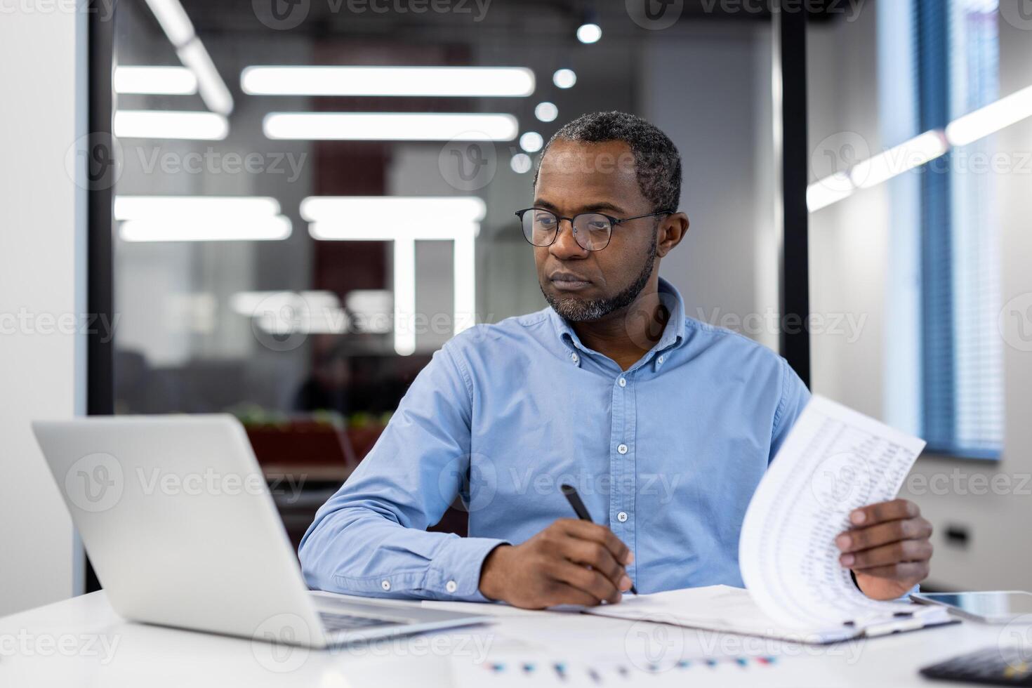 Mature African American businessman in modern office working on laptop, reviewing documents. Focused, professional, productive atmosphere, emphasizing business analysis, decision-making, and strategy photo