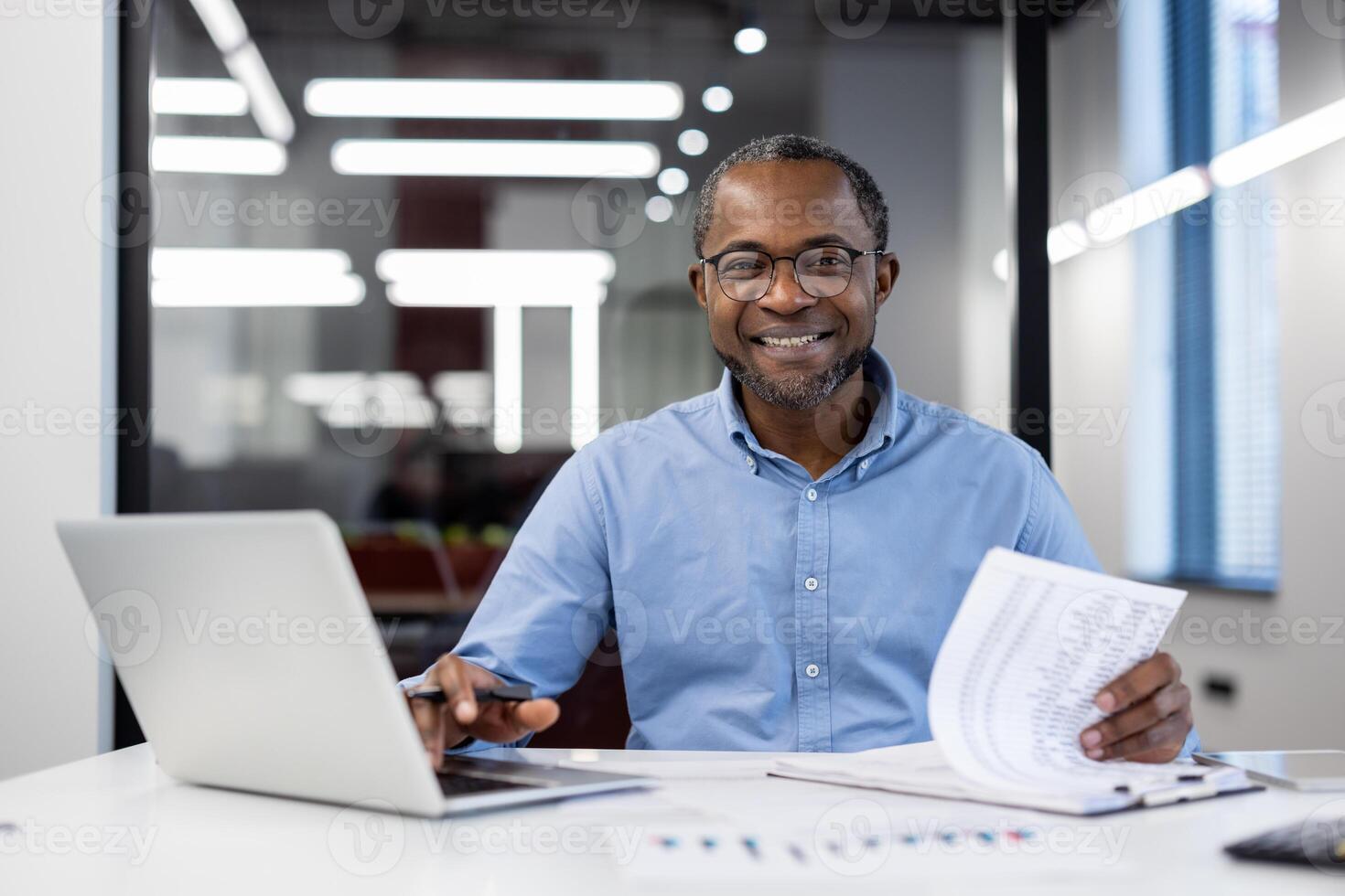 Mature african american businessman smiling confidently working on laptop in modern office. Business setting with important documents showcasing success and professionalism. photo