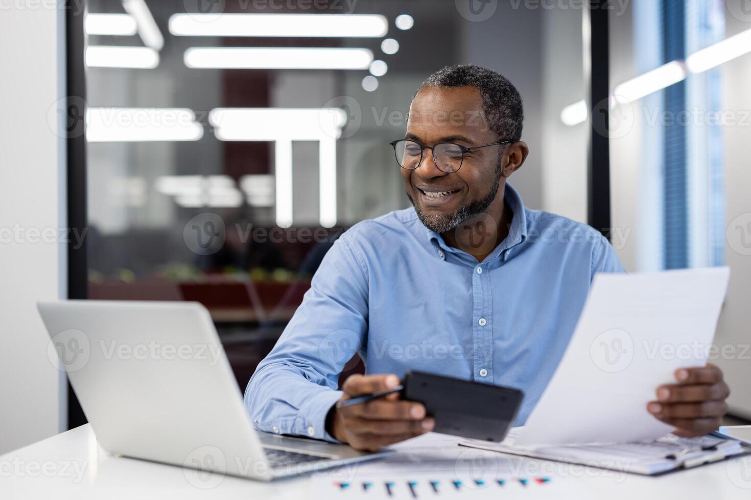 Mature African American businessman smiling while reviewing documents in modern office setting with laptop and smartphone. Professional environment emphasizes productivity, achievement, success. photo