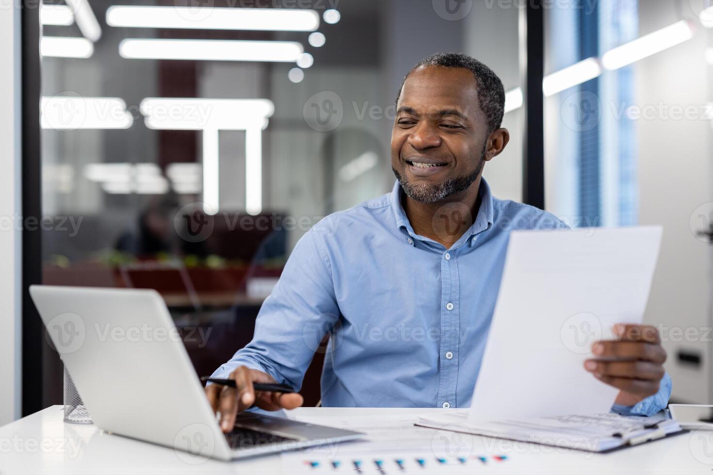 Mature African American businessman reviewing business documents at desk using laptop in modern office setting. Confident, focused, smiling professional in blue shirt during work session. photo