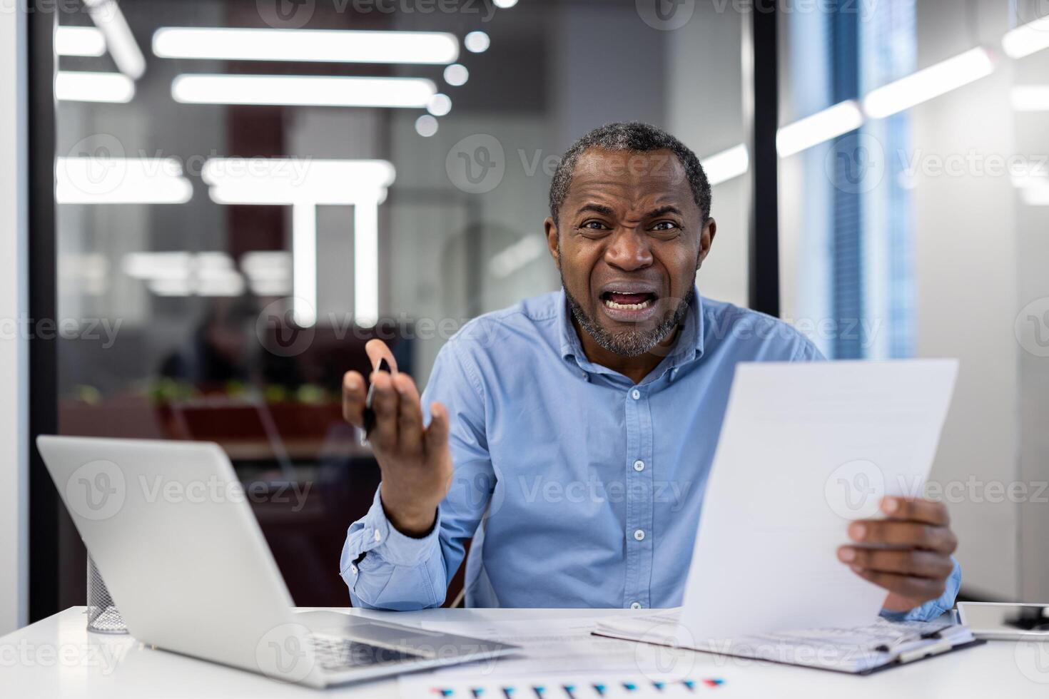 African American businessman in office showing frustration with document and laptop. Business environment highlights stress, challenges. Concept pressure, communication, emotion in modern workplace. photo