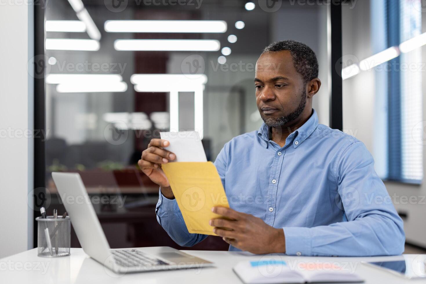 Mature African American businessman in office using laptop, opening envelope. Focused expression suggests concentration, professionalism. Bright lighting, modern setting denotes corporate environment photo