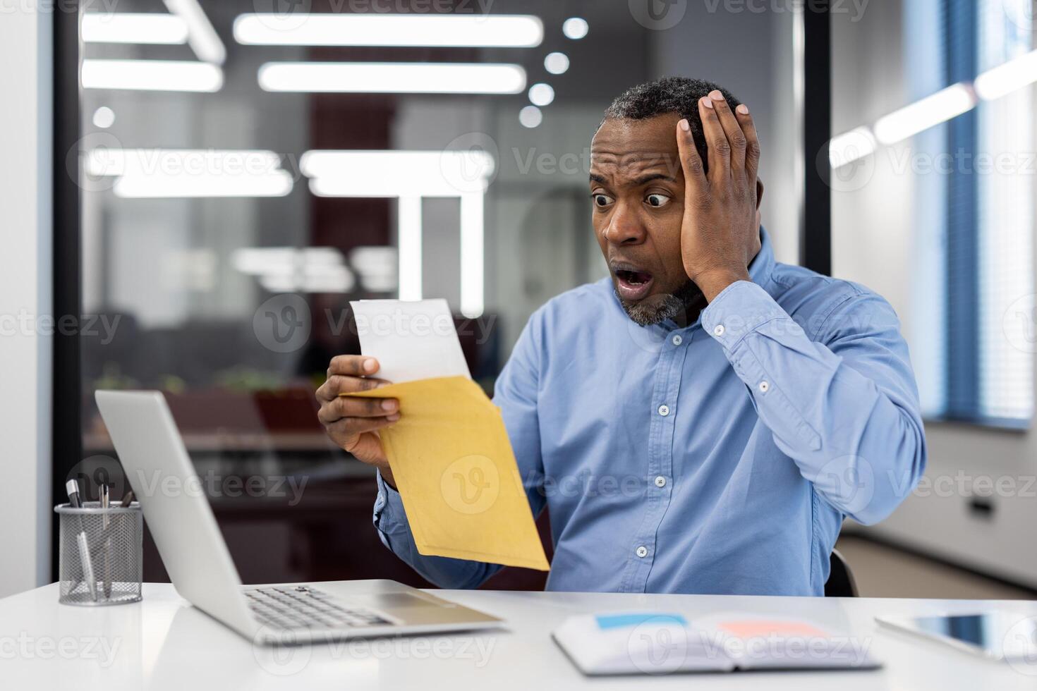 Mature African American businessman looks shocked while reading surprising news at office desk. Open mouth and hand-on-face reveal intense reaction. Laptop and document illustrate work environment. photo