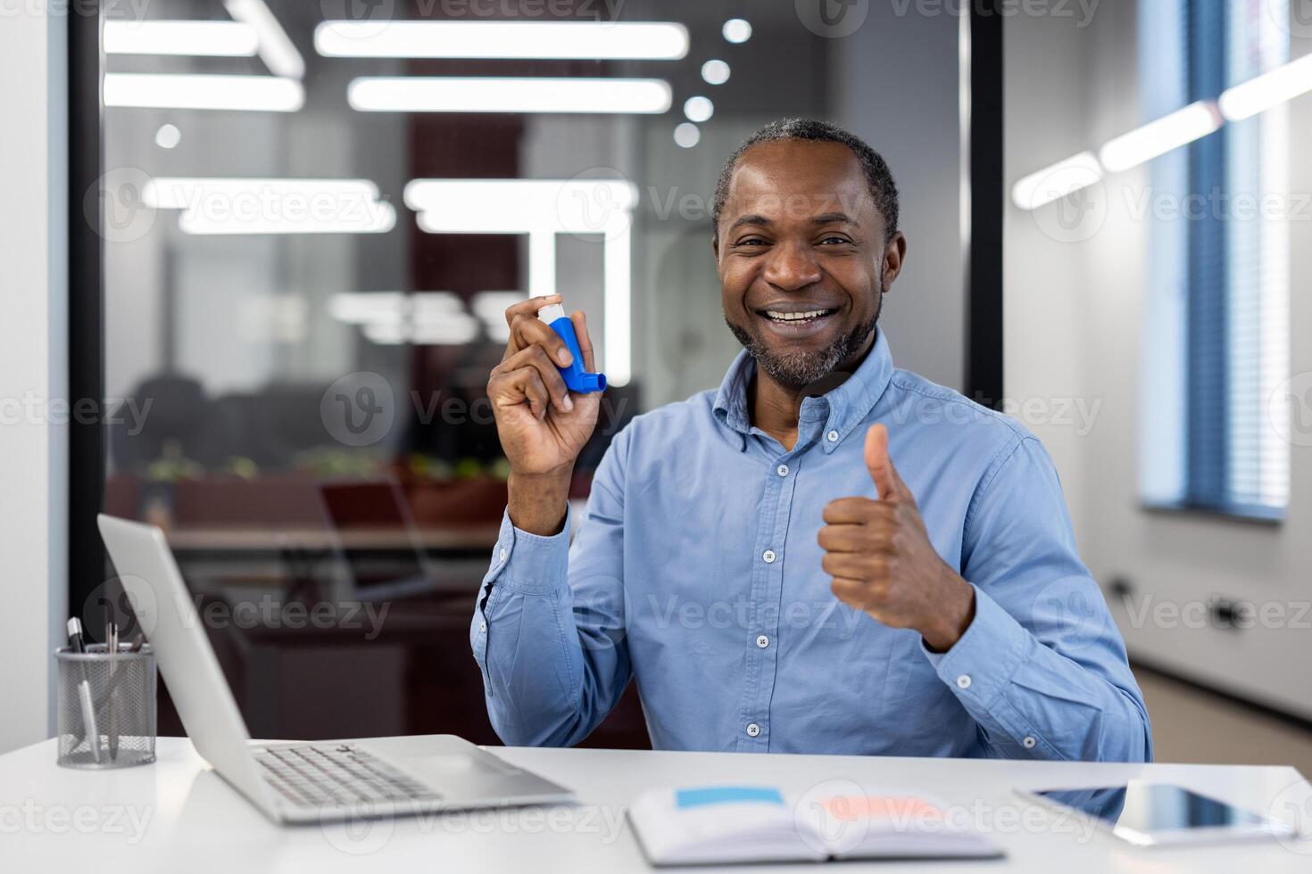 Mature businessman smiling in modern office, holding asthma inhaler, showing thumbs up. Represents health management, positive attitude, success in workplace wellness. photo