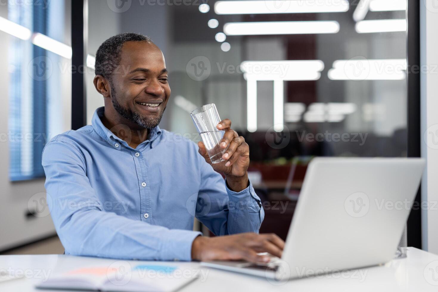 Mature African American businessman smiling while working on laptop in modern office. Holding glass of water, showing positive and professional attitude. Concept of success and productivity. photo