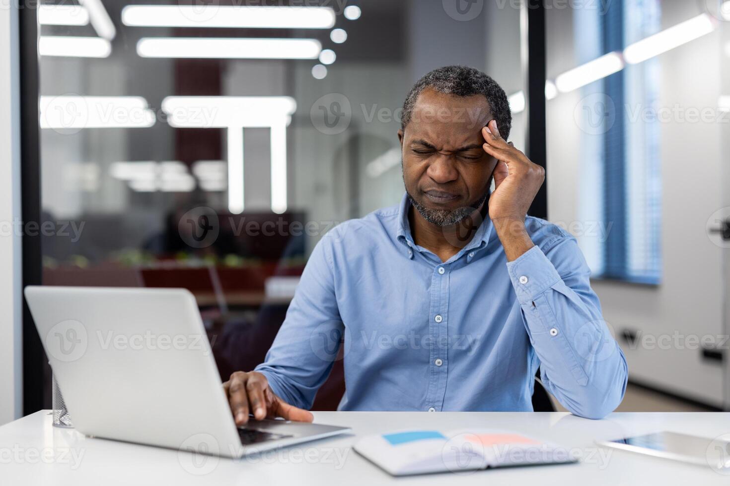Mature African American businessman experiences stress at office desk, working on laptop. Hand on temple shows tension. Professional environment suggests busy workday. photo