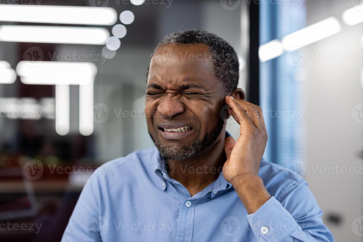 Mature African American businessman in modern office setting, showing discomfort by holding ear and wincing. Expression of frustration, possibly due to noise or stress-related issue. photo