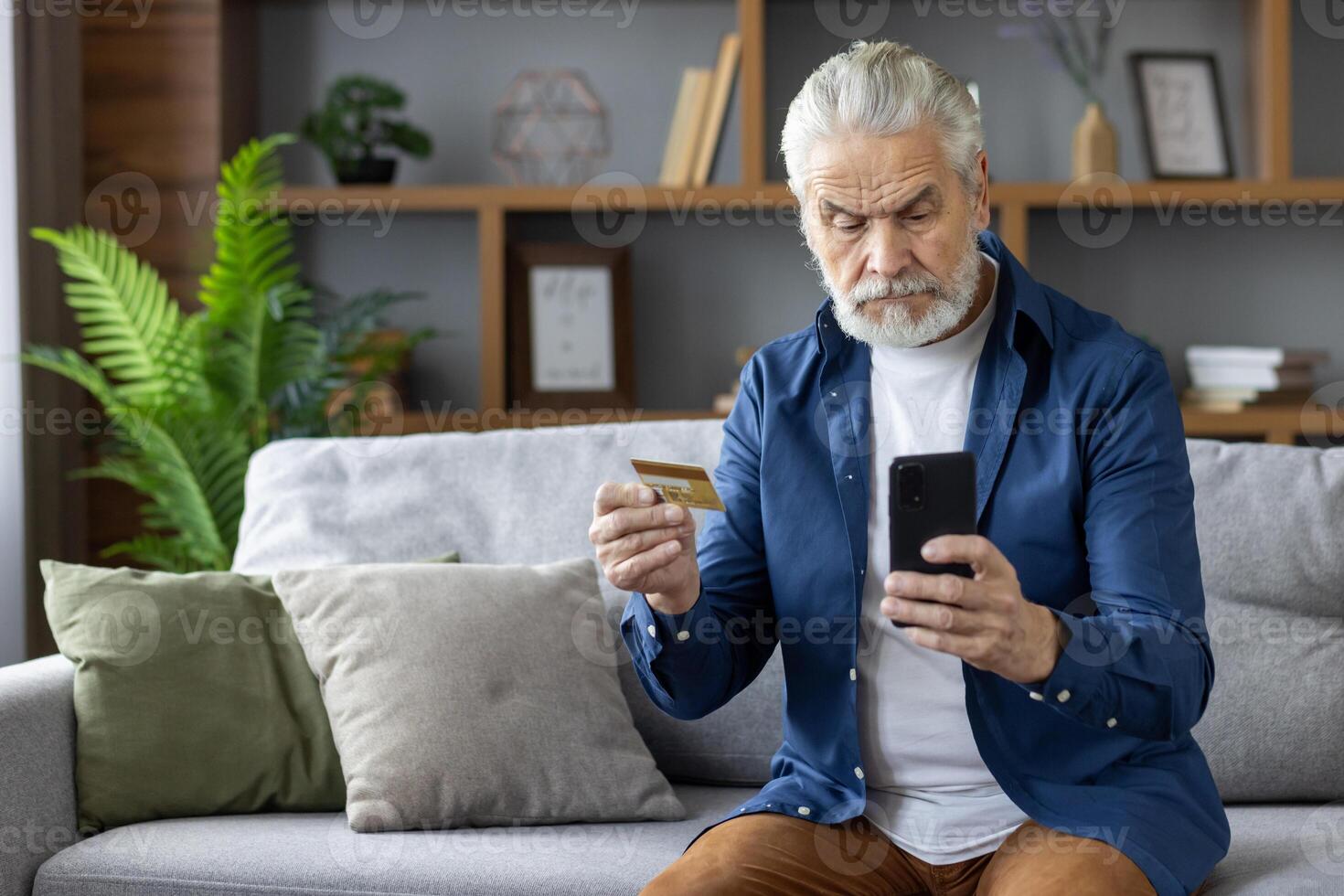 A senior man looks puzzled as he holds a credit card and uses a smartphone in a modern living room. The concept of technology challenges for the elderly. photo