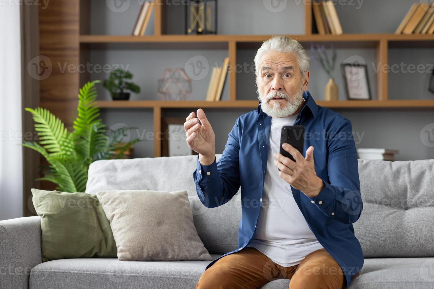 Elderly Caucasian man looking puzzled and stressed while looking at his mobile phone, sitting on a sofa in a well-lit living room. photo