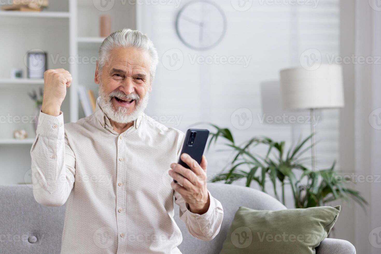 Happy gray-haired senior man is at home, sitting on the couch, looking at the screen of the phone he is holding and rejoicing in success, showing a victory gesture with his hand. photo