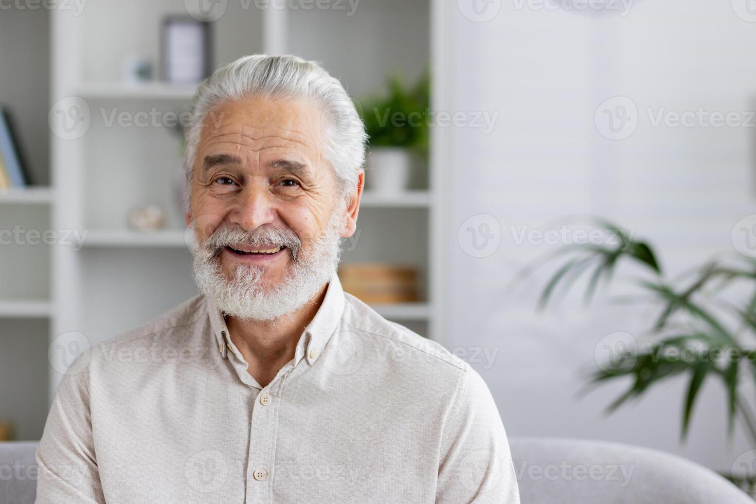 Close-up portrait of a smiling gray-haired senior man at home, sitting on a sofa in a white shirt and with a beard, smiling at the camera. photo